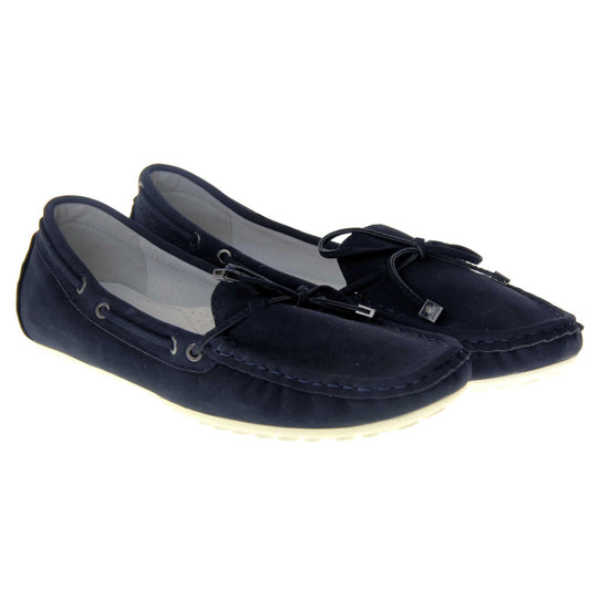 Women's deck shoes. Moccasin style boat shoes with a navy blue faux suede upper. Stitching detail around the top and a lace and bow detail around the collar and tongue of the shoe. Grey leather lining and thin white sole. Both feet together from an angle.