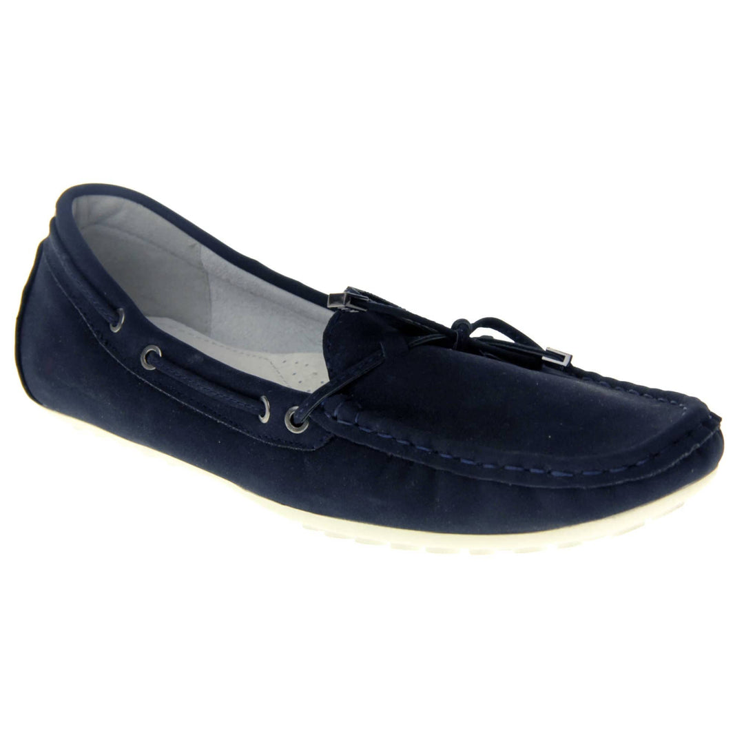 Women's deck shoes. Moccasin style boat shoes with a navy blue faux suede upper. Stitching detail around the top and a lace and bow detail around the collar and tongue of the shoe. Grey leather lining and thin white sole. Right foot at an angle.
