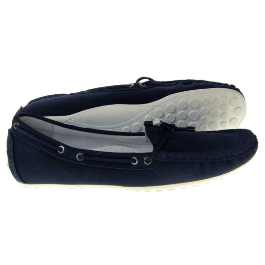 Women's deck shoes. Moccasin style boat shoes with a navy blue faux suede upper. Stitching detail around the top and a lace and bow detail around the collar and tongue of the shoe. Grey leather lining and thin white sole. Both feet from a side profile with the left foot on its side to show the sole.