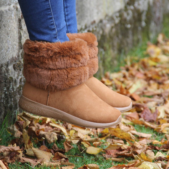 Womens Faux Fur Lined Winter Boots - Tan Brown with slight wedge heel, soft faux fur cuff. Model shot in Autumn, surrounded by leaves and beside a wall.