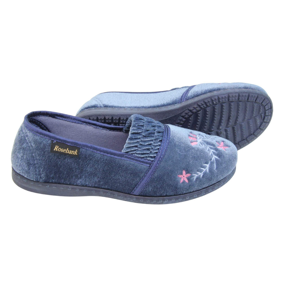 Womens full slippers. Full back slippers in a loafer style. With light blue velour uppers and an embroidered pale blue and pink flower detail. Ruched velour elasticated gusset. Blue textile lining and piping around the collar. Blue firm sole. Both feet from a side profile with the left foot on its side behind the the right foot to show the sole.