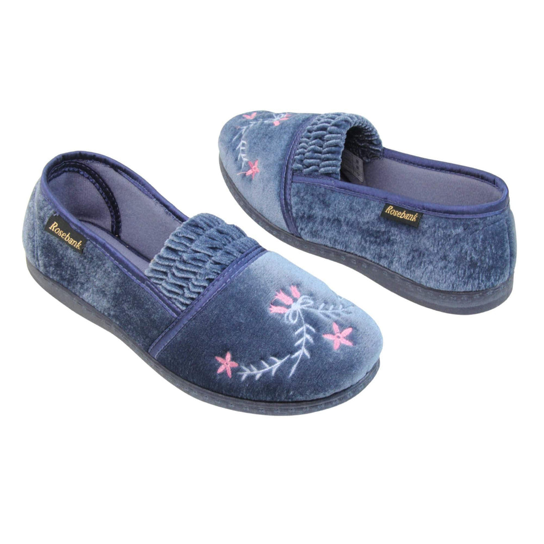 Womens full slippers. Full back slippers in a loafer style. With light blue velour uppers and an embroidered pale blue and pink flower detail. Ruched velour elasticated gusset. Blue textile lining and piping around the collar. Blue firm sole. Both feet at an angle facing top to tail.