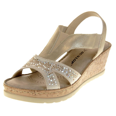 Womens gold wedge sandals. Pale gold coloured, faux leather upper. Diamantes along the toe straps. Elasticated panels in the middle of the central strap and where the heel strap meets the central strap. Nude insole with black Dunlop branding. Cork heel and platform with beige outsole with tread to the bottom. Left foot at an angle.