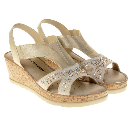 Womens gold wedge sandals. Pale gold coloured, faux leather upper. Diamantes along the toe straps. Elasticated panels in the middle of the central strap and where the heel strap meets the central strap. Nude insole with black Dunlop branding. Cork heel and platform with beige outsole with tread to the bottom. Both feet together at a slight angle.