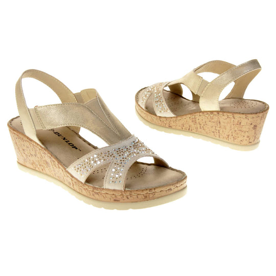 Womens gold wedge sandals. Pale gold coloured, faux leather upper. Diamantes along the toe straps. Elasticated panels in the middle of the central strap and where the heel strap meets the central strap. Nude insole with black Dunlop branding. Cork heel and platform with beige outsole with tread to the bottom. Both feet at an angle facing top to tail.