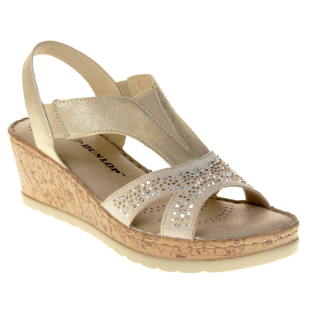 Womens gold wedge sandals. Pale gold coloured, faux leather upper. Diamantes along the toe straps. Elasticated panels in the middle of the central strap and where the heel strap meets the central strap. Nude insole with black Dunlop branding. Cork heel and platform with beige outsole with tread to the bottom. Right foot at an angle.