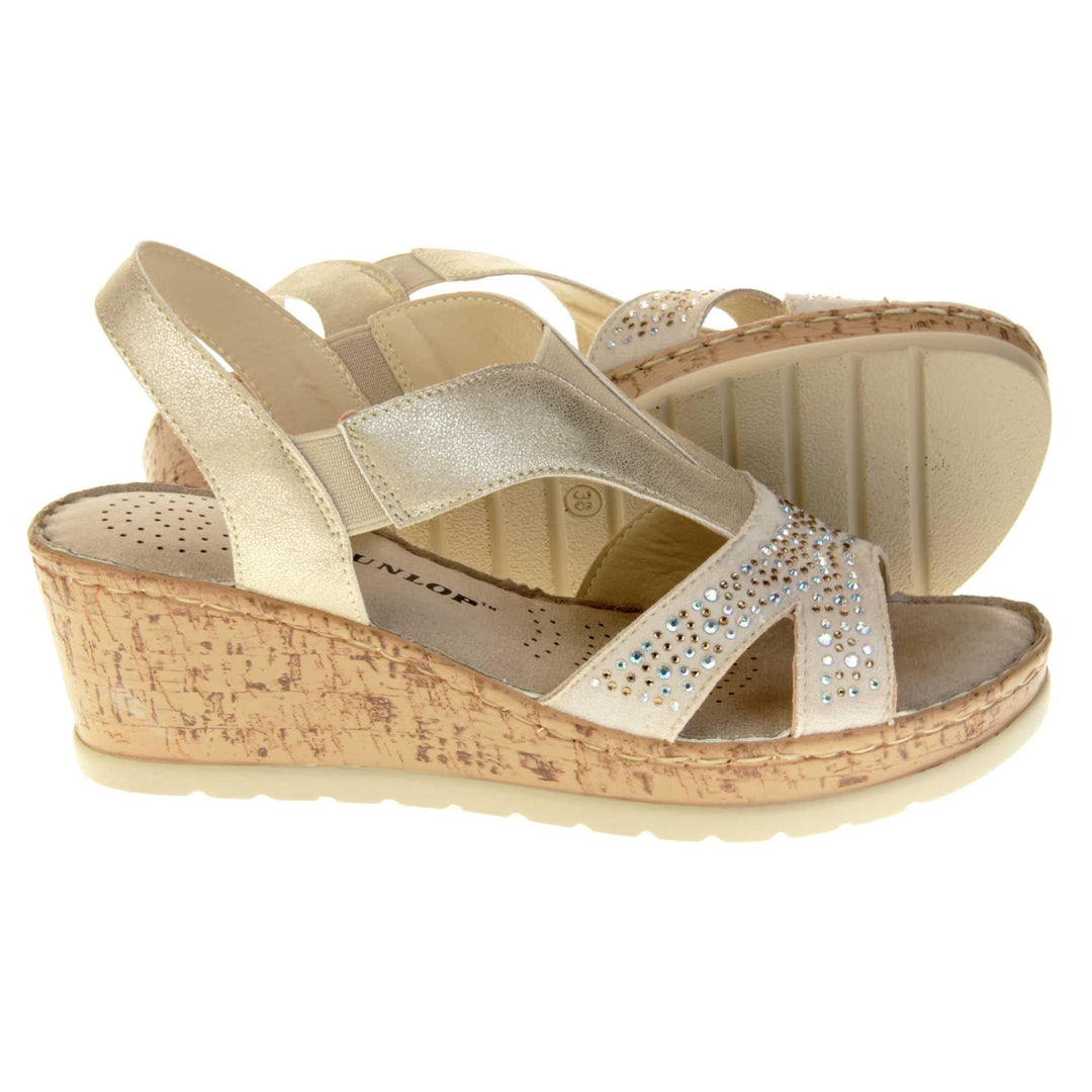 Womens gold wedge sandals. Pale gold coloured, faux leather upper. Diamantes along the toe straps. Elasticated panels in the middle of the central strap and where the heel strap meets the central strap. Nude insole with black Dunlop branding. Cork heel and platform with beige outsole with tread to the bottom. Both feet from a side profile with the left foot on its side behind the the right foot to show the sole.