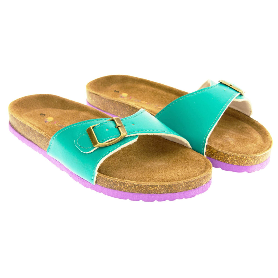 Womens green sliders. Green faux leather strap with gold buckle. Soft tan faux suede footbed with cork effect outsole and purple sole. Both feet together at a slight angle.