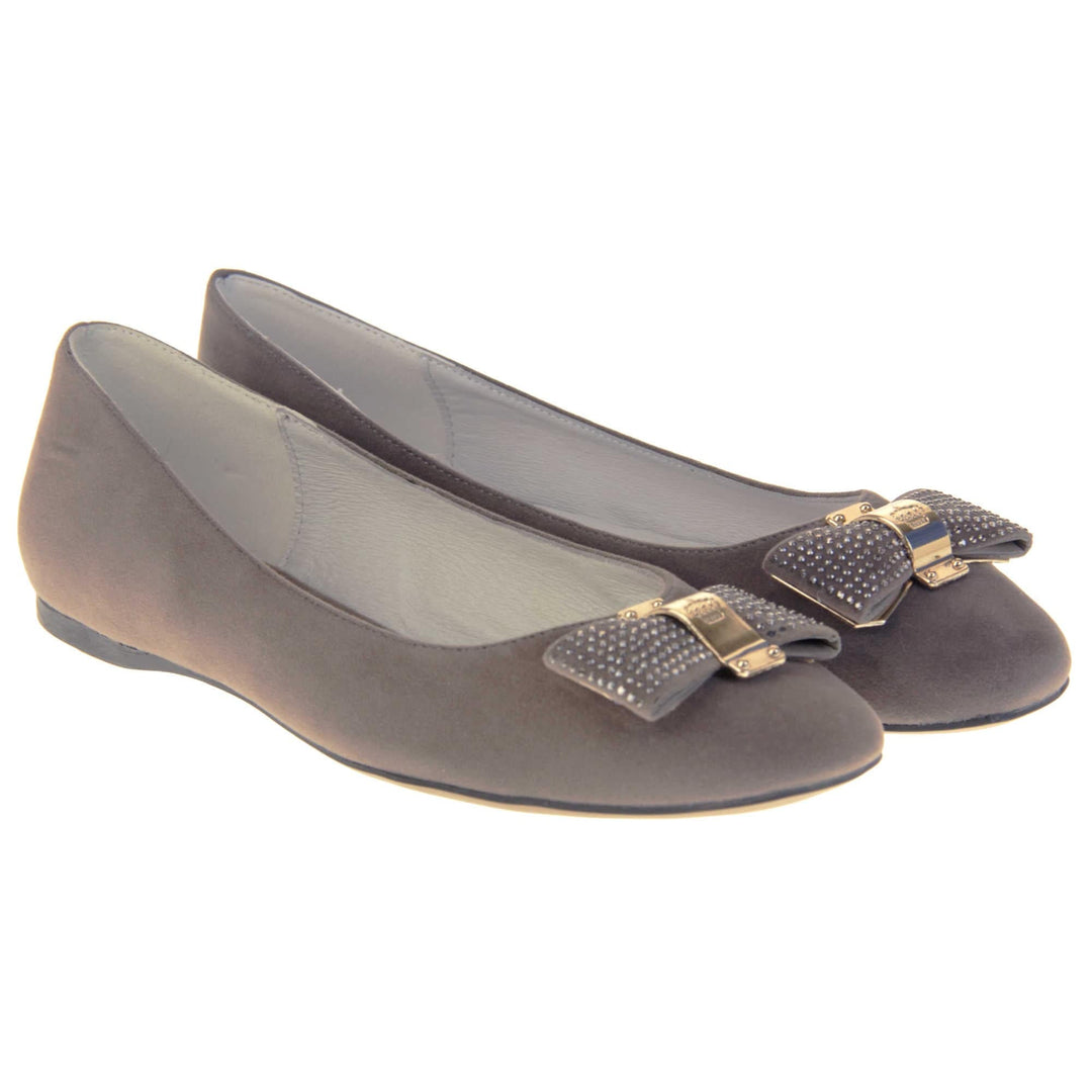 Women's grey flats. Ballet style shoes with a grey faux suede upper. Grey bow detail to the top with diamantes covering it and a gold coloured metal loop round the middle. Cream leather lining and black sole. Both feet together at a slight angle.