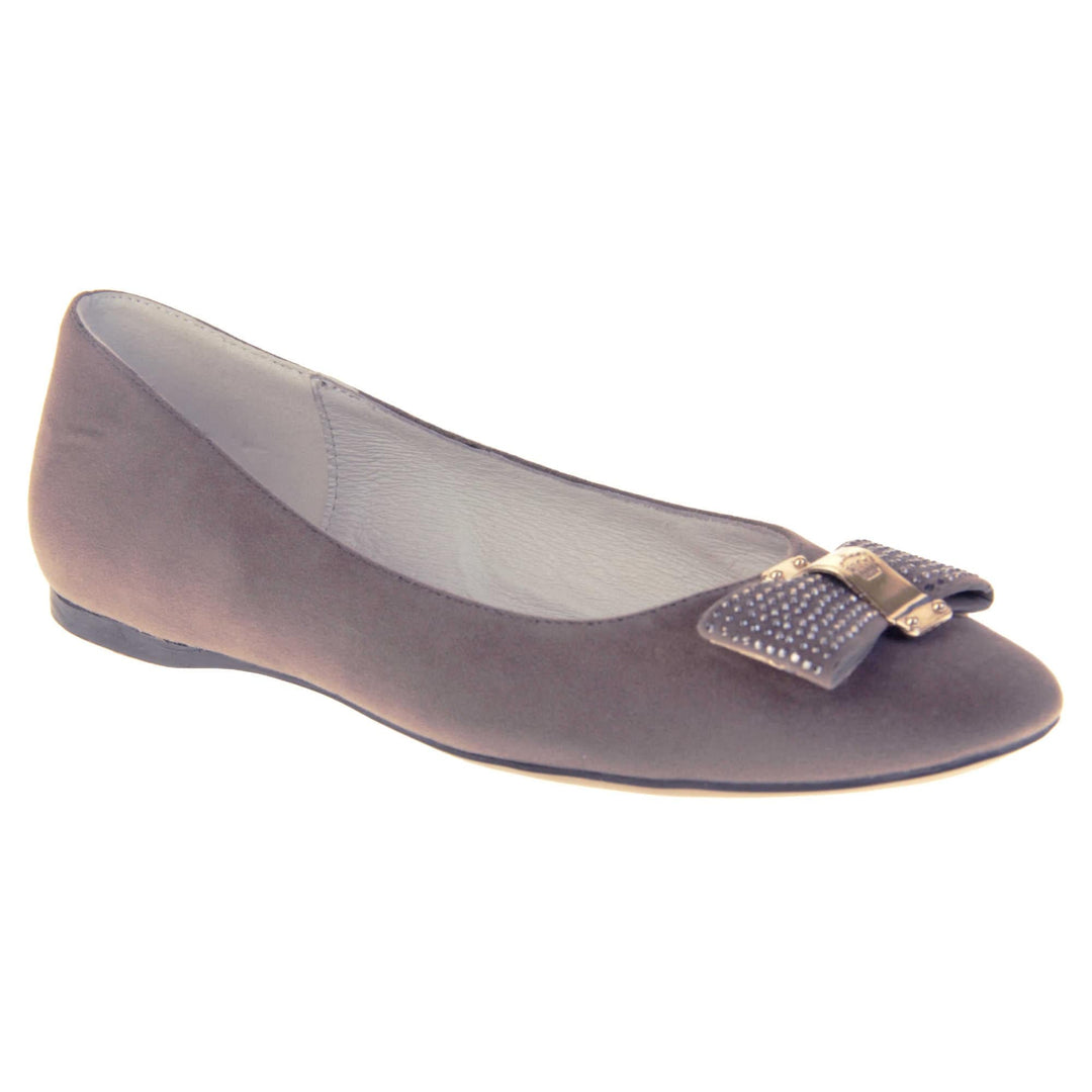 Women's grey flats. Ballet style shoes with a grey faux suede upper. Grey bow detail to the top with diamantes covering it and a gold coloured metal loop round the middle. Cream leather lining and black sole. Right foot at an angle.