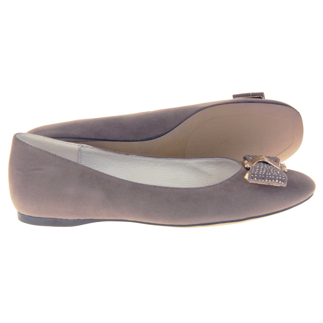 Women's grey flats. Ballet style shoes with a grey faux suede upper. Grey bow detail to the top with diamantes covering it and a gold coloured metal loop round the middle. Cream leather lining and black sole.  Both feet from a side profile with the left foot on its side behind the the right foot to show the sole.