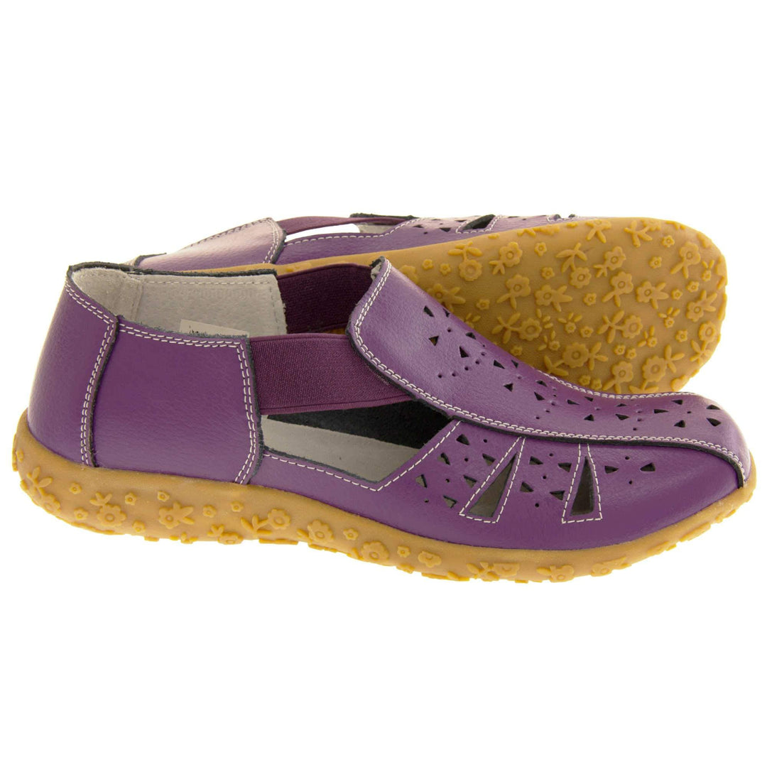 Womens leather sandals. Purple leather closed toe sandals with white stitched detailing. With small cut out details on the upper. Purple elasticated strips from tongue to ankle to allow more room for a better fit. Cream coloured leather insole and lining. Brown sole with heel having a slight platform with raised flower design for grip. Left foot at an angle. Both feet from side profile with left foot on its side behind the right to show the sole.