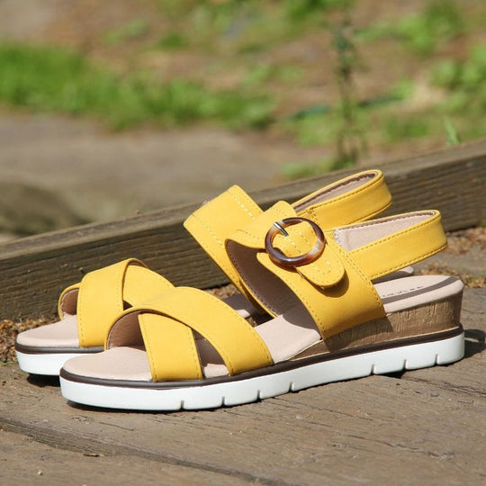 Womens Low Wedge Sandals - Ivory Castle Mustard yellow faux leather upper with touch fastening strap over. Memory foam insoles and white flexible outsole. Brown decorative buckle to side. Crossover straps to font. Classic timeless style. Side shot on bridge in park.