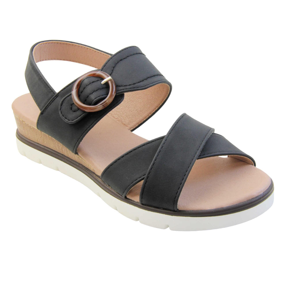 Womens memory foam sandals. Classic womens strappy sandals with black textile straps. Dual toe straps that cross over each other. The ankle strap is touch fasten but has a brown buckle detail to look like a buckle fastening. Beige faux leather memory foam insoles. Small wedge heel in cork effect. White outsole with a black rim around the top. Right foot at an angle.