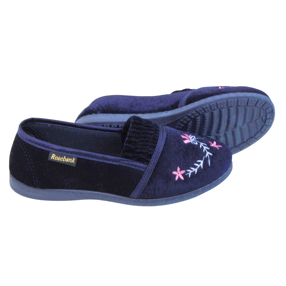 Womens navy slippers. Full back slippers in a loafer style. With navy blue velour uppers and a embroidered pale blue and pink flower detail. Ruched velour elasticated gusset. Navy textile lining and piping around the collar. Dark blue firm sole. Both feet from a side profile with the left foot on its side behind the the right foot to show the sole.