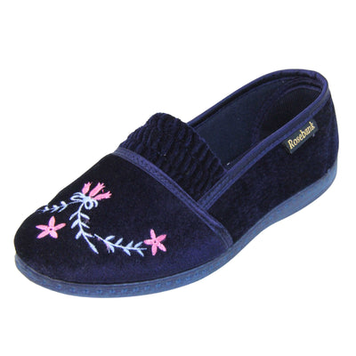 Womens navy slippers. Full back slippers in a loafer style. With navy blue velour uppers and a embroidered pale blue and pink flower detail. Ruched velour elasticated gusset. Navy textile lining and piping around the collar. Dark blue firm sole. Left foot at an angle.