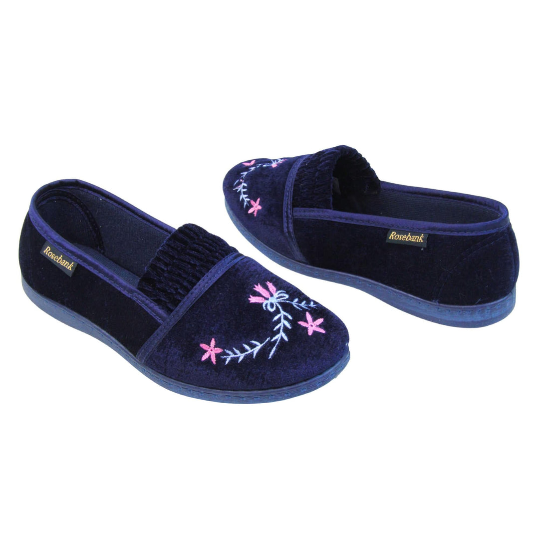 Womens navy slippers. Full back slippers in a loafer style. With navy blue velour uppers and a embroidered pale blue and pink flower detail. Ruched velour elasticated gusset. Navy textile lining and piping around the collar. Dark blue firm sole. Both feet at an angle facing top to tail.