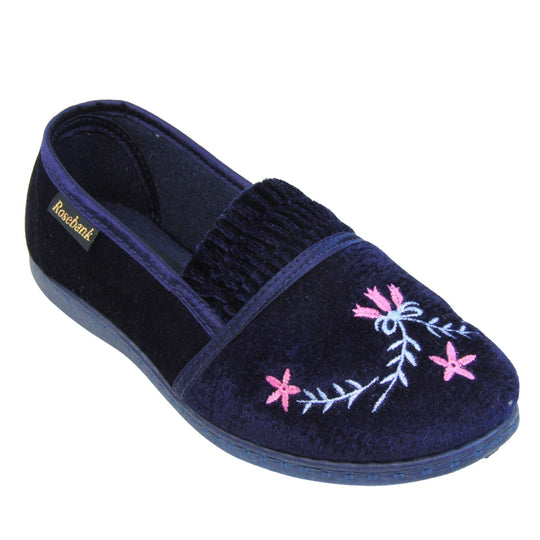 Womens navy slippers. Full back slippers in a loafer style. With navy blue velour uppers and a embroidered pale blue and pink flower detail. Ruched velour elasticated gusset. Navy textile lining and piping around the collar. Dark blue firm sole. Right foot at an angle.