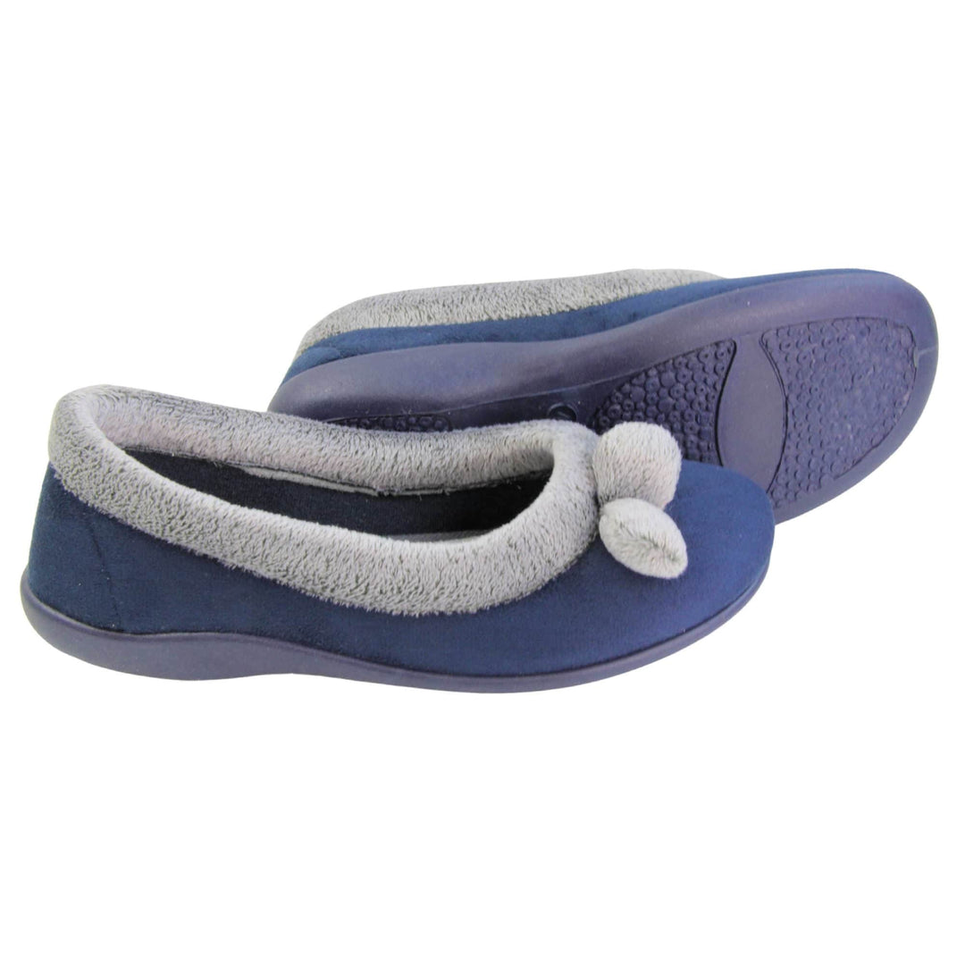 Womens navy slippers. Womens ballerina style slippers with navy blue velour uppers. Grey, plush textile collar and two pom poms to the front of the shoe. Matching textile lining. Firm navy sole. Both feet from a side profile with the left foot on its side behind the the right foot to show the sole.