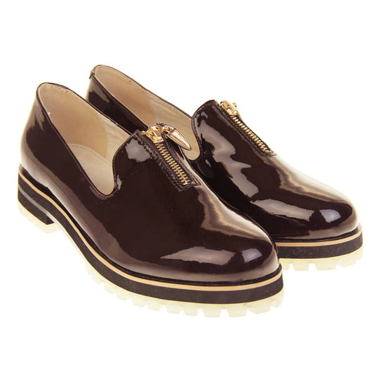 Womens red loafers. Loafer style shoes with dark burgundy patent uppers. Short gold zip detail to the top of the shoe. Black sole with cream base and small heel and beige leather lining. Both feet together at an angle.