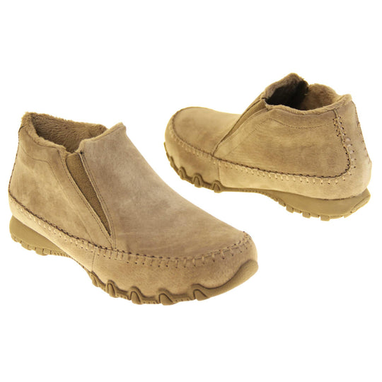 Womens Skechers boots. Sand coloured suede uppers in an ankle boot style. With brown elasticated panels by the tongue. Brown chenille lining. Brown sole with grip the bottom. Both feet at a slight angle facing top to tail.
