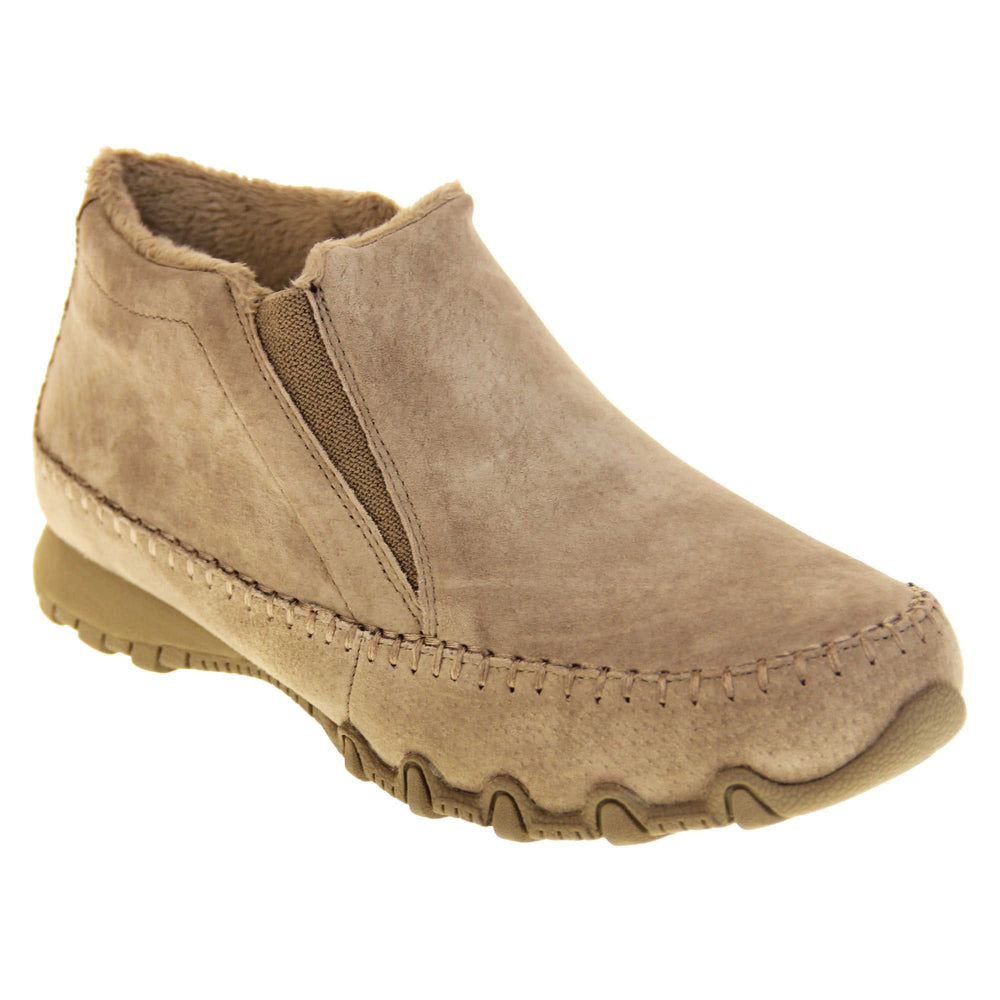 Womens Skechers boots. Sand coloured suede uppers in an ankle boot style. With brown elasticated panels by the tongue. Brown chenille lining. Brown sole with grip the bottom. Right foot at an angle.