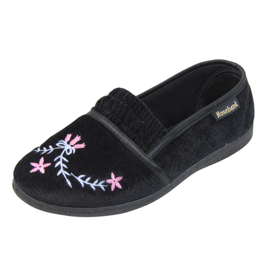 Womens soft slippers. Full back slippers in a loafer style. With black velour uppers and an embroidered pale blue and pink flower detail. Ruched velour elasticated gusset. Black textile lining and piping around the collar. Black firm sole. Left foot at an angle.