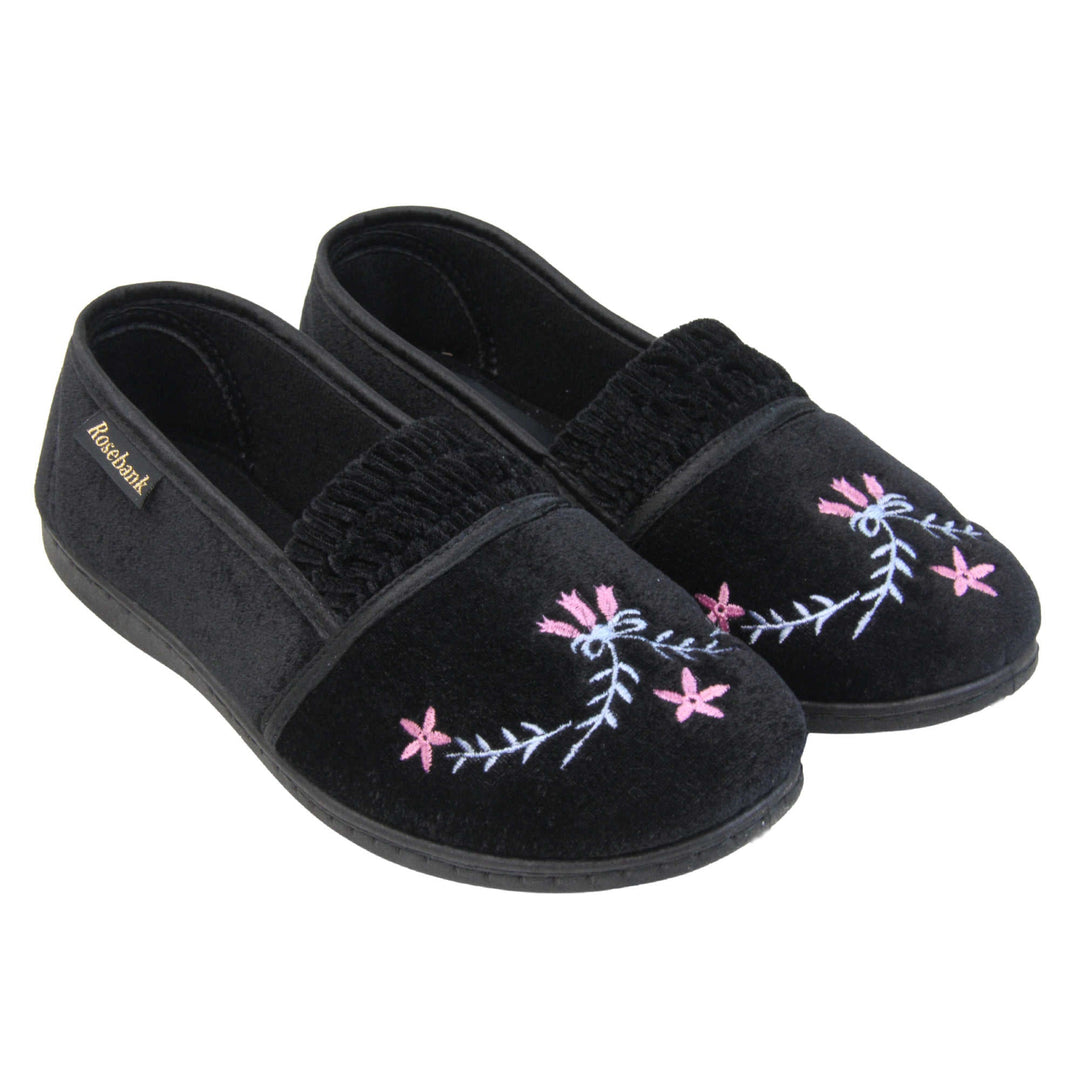Womens soft slippers. Full back slippers in a loafer style. With black velour uppers and an embroidered pale blue and pink flower detail. Ruched velour elasticated gusset. Black textile lining and piping around the collar. Black firm sole. Both feet together at an angle.