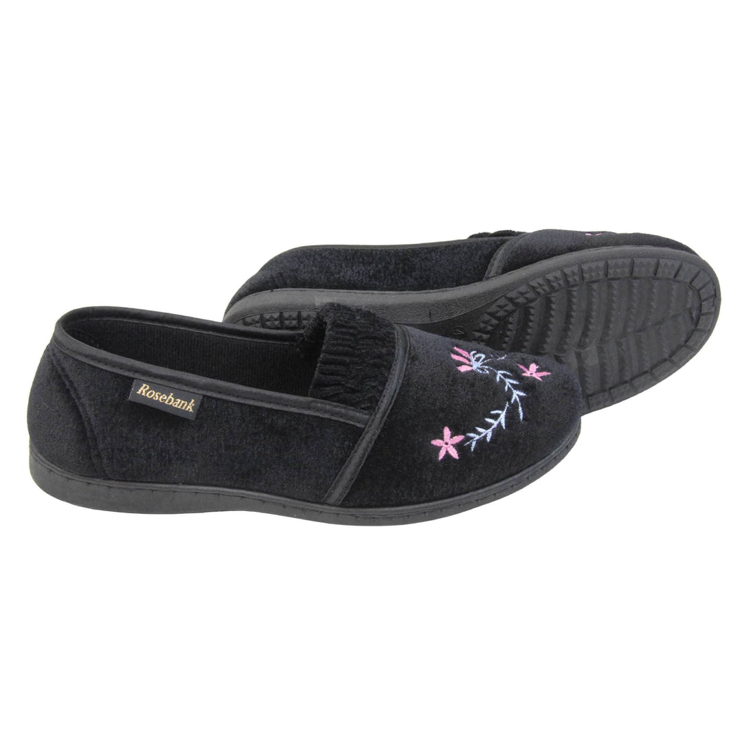 Womens soft slippers. Full back slippers in a loafer style. With black velour uppers and an embroidered pale blue and pink flower detail. Ruched velour elasticated gusset. Black textile lining and piping around the collar. Black firm sole. Both feet from a side profile with the left foot on its side behind the the right foot to show the sole.