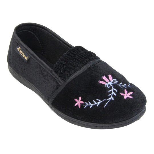Womens soft slippers. Full back slippers in a loafer style. With black velour uppers and an embroidered pale blue and pink flower detail. Ruched velour elasticated gusset. Black textile lining and piping around the collar. Black firm sole. Right foot at an angle.