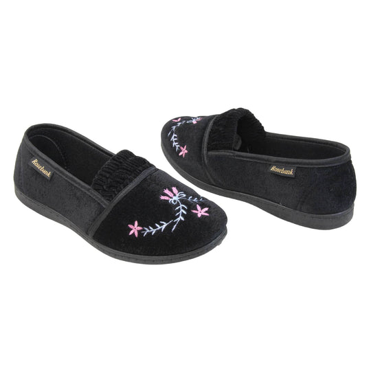 Womens soft slippers. Full back slippers in a loafer style. With black velour uppers and an embroidered pale blue and pink flower detail. Ruched velour elasticated gusset. Black textile lining and piping around the collar. Black firm sole. Both feet at an angle facing top to tail.