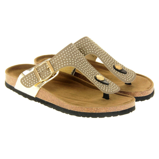Womens studded sandals. Taupe faux leather strap covered in gold studs. With black toe post to the front and gold buckle to the outside. Metallic gold strap to the side meeting at the buckle. Soft tan faux suede footbed with cork effect outsole and black sole. Both feet together at a slight angle.