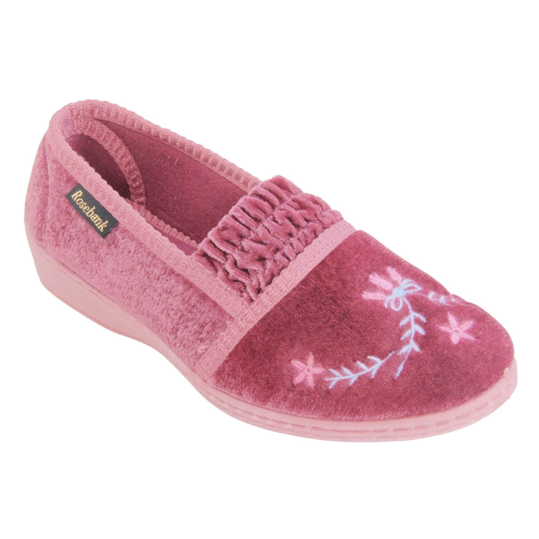 Womens wedge slippers. Full back slippers in a loafer style. With pink velour uppers and a embroidered pale blue and pink flower detail. Ruched velour elasticated gusset. Pink textile lining and piping around the collar. Pink firm sole with a small wedge heel. Right foot at an angle.