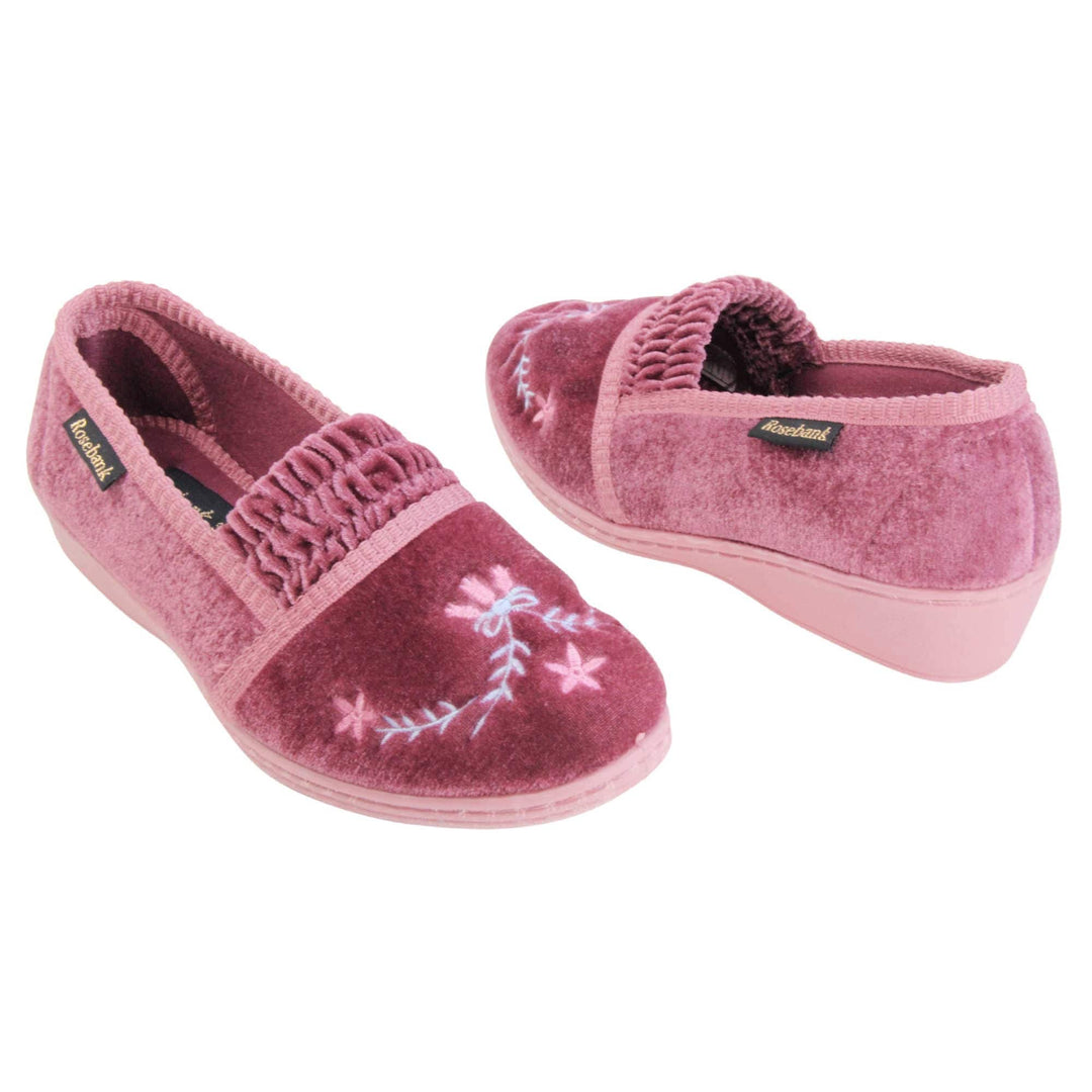 Womens wedge slippers. Full back slippers in a loafer style. With pink velour uppers and a embroidered pale blue and pink flower detail. Ruched velour elasticated gusset. Pink textile lining and piping around the collar. Pink firm sole with a small wedge heel. Both feet at an angle facing top to tail.