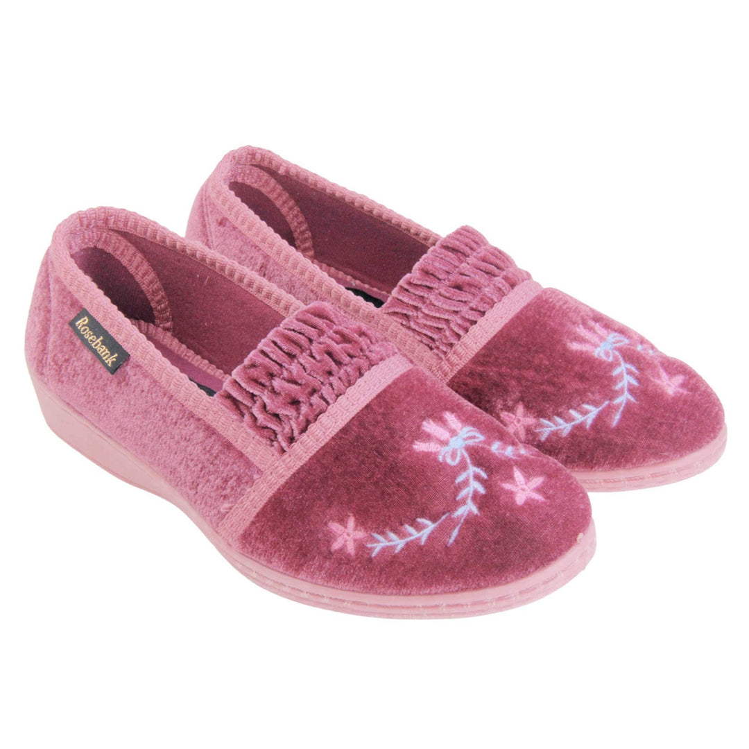 Womens wedge slippers. Full back slippers in a loafer style. With pink velour uppers and a embroidered pale blue and pink flower detail. Ruched velour elasticated gusset. Pink textile lining and piping around the collar. Pink firm sole with a small wedge heel. Both feet together at an angle.