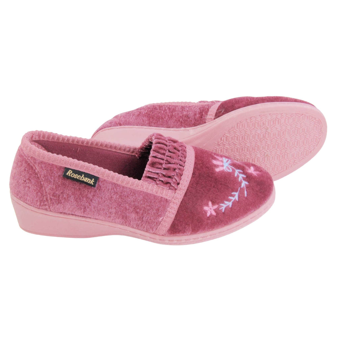 Womens wedge slippers. Full back slippers in a loafer style. With pink velour uppers and a embroidered pale blue and pink flower detail. Ruched velour elasticated gusset. Pink textile lining and piping around the collar. Pink firm sole with a small wedge heel. Both feet from a side profile with the left foot on its side behind the the right foot to show the sole.