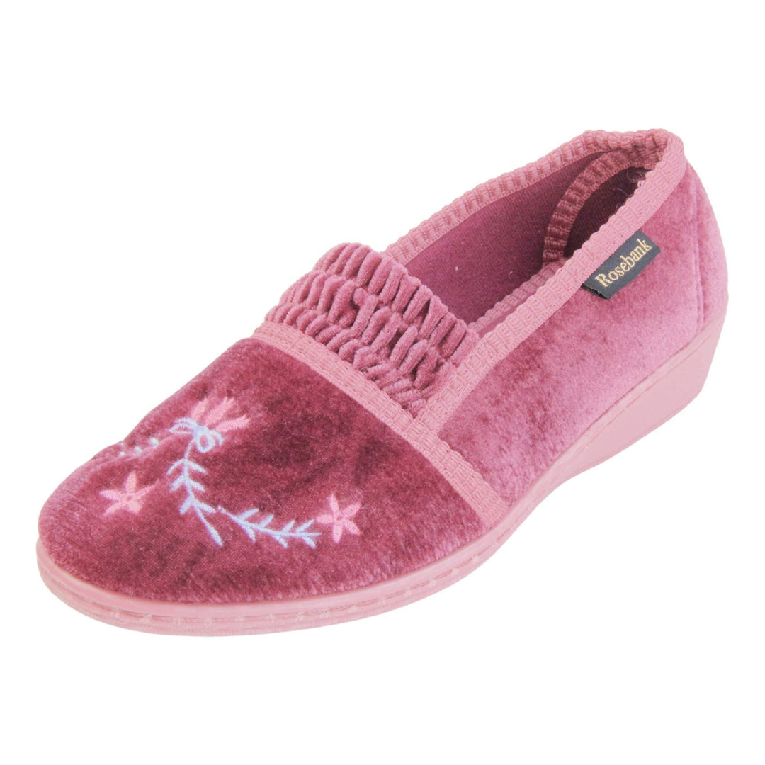Womens wedge slippers. Full back slippers in a loafer style. With pink velour uppers and a embroidered pale blue and pink flower detail. Ruched velour elasticated gusset. Pink textile lining and piping around the collar. Pink firm sole with a small wedge heel. Left foot at an angle.