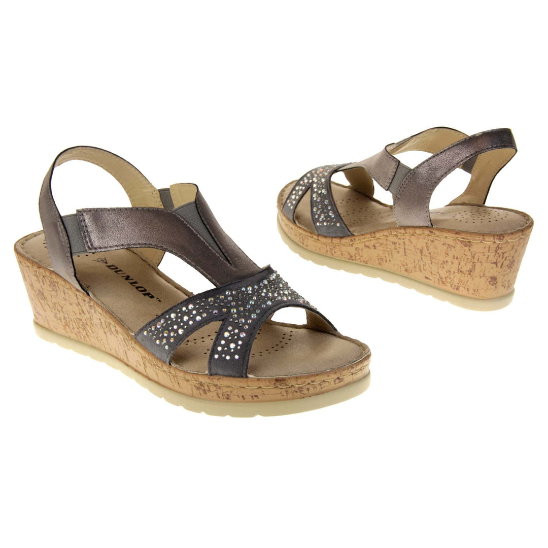 Womens wedge sandals. Pewter coloured, faux leather upper. Diamantes along the toe straps. Elasticated panels in the middle of the central strap and where the heel strap meets the central strap. Brown insole with black Dunlop branding. Cork heel and platform with beige outsole with tread to the bottom. Both feet at an angle facing top to tail.