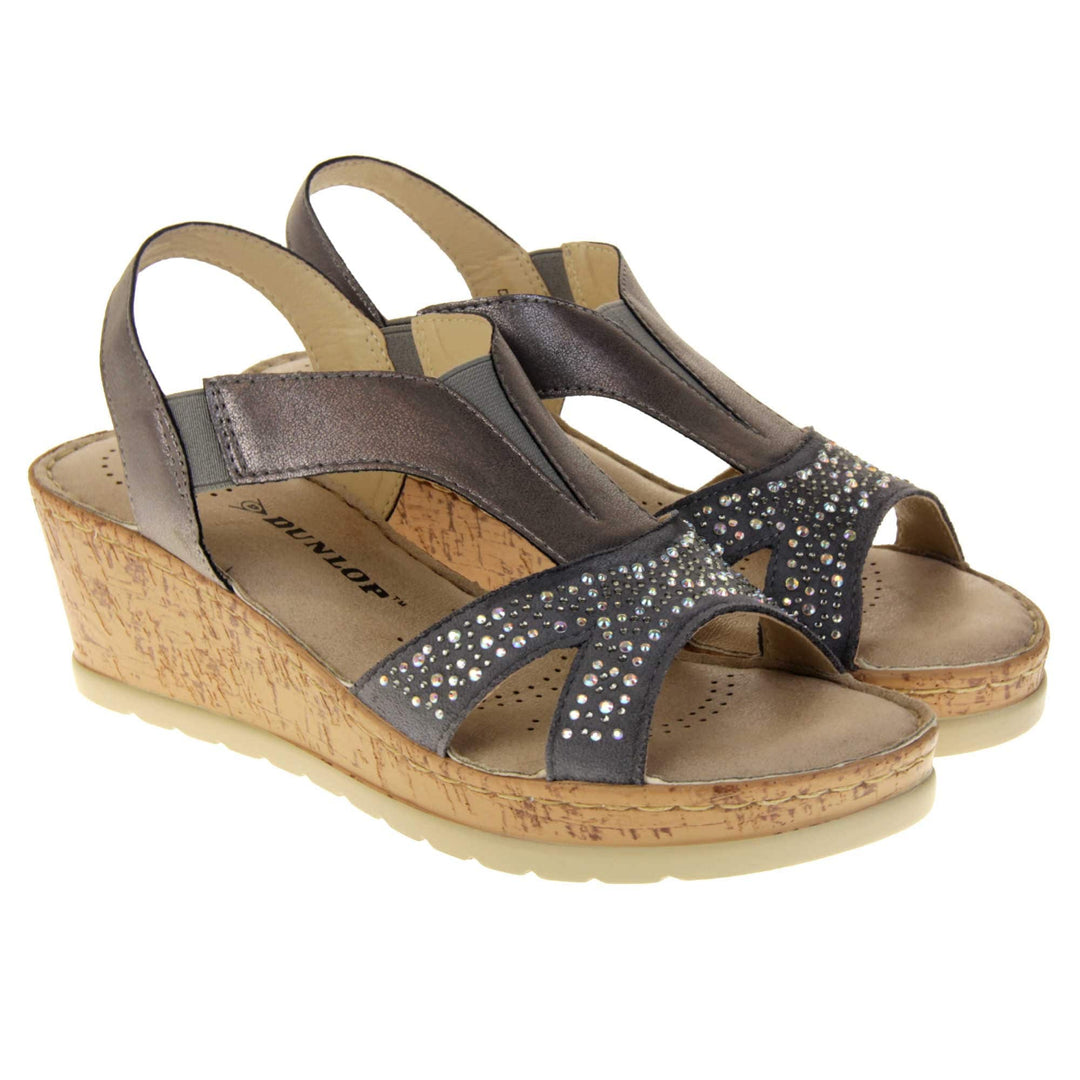 Womens wedge sandals. Pewter coloured, faux leather upper. Diamantes along the toe straps. Elasticated panels in the middle of the central strap and where the heel strap meets the central strap. Brown insole with black Dunlop branding. Cork heel and platform with beige outsole with tread to the bottom. Both feet together at a slight angle.