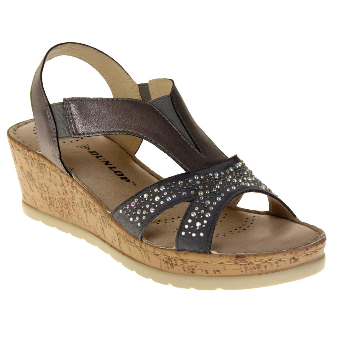 Womens wedge sandals. Pewter coloured, faux leather upper. Diamantes along the toe straps. Elasticated panels in the middle of the central strap and where the heel strap meets the central strap. Brown insole with black Dunlop branding. Cork heel and platform with beige outsole with tread to the bottom. Right foot at an angle.