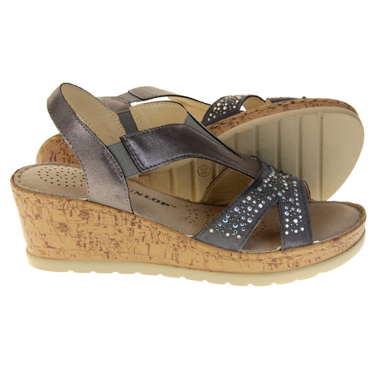 Womens wedge sandals. Pewter coloured, faux leather upper. Diamantes along the toe straps. Elasticated panels in the middle of the central strap and where the heel strap meets the central strap. Brown insole with black Dunlop branding. Cork heel and platform with beige outsole with tread to the bottom.  Both feet from a side profile with the left foot on its side behind the the right foot to show the sole.
