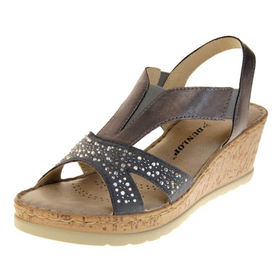 Womens wedge sandals. Pewter coloured, faux leather upper. Diamantes along the toe straps. Elasticated panels in the middle of the central strap and where the heel strap meets the central strap. Brown insole with black Dunlop branding. Cork heel and platform with beige outsole with tread to the bottom. Left foot at an angle.