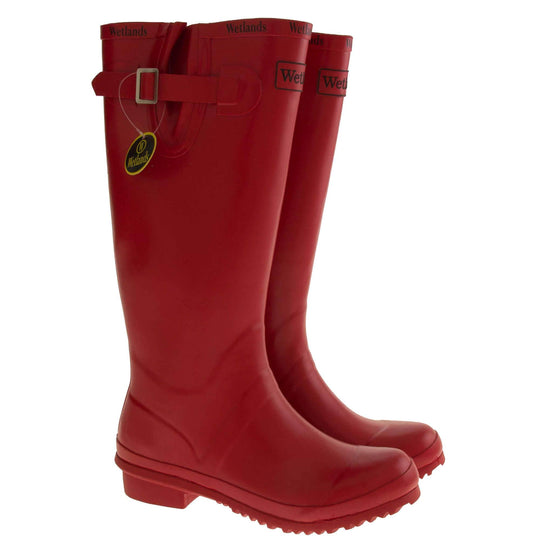 Womens Wellies. Red rubber waterproof wellington boots. Just below knee height. With a small heel and deep tread to the sole. Black Wetlands Brand to the front of the boot near the top. Wetlands written in black numerous times around the rim of the boot. Side strap with buckle to adjust the calf width. Textile lining to the inside of the boot. Both shoes next to each other from slight side angle