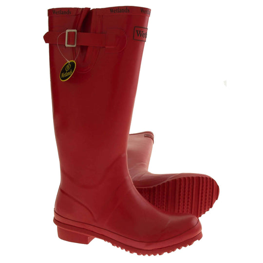 Womens Wellies. Red rubber waterproof wellington boots. Just below knee height. With a small heel and deep tread to the sole. Black Wetlands Brand to the front of the boot near the top. Wetlands written in black numerous times around the rim of the boot. Side strap with buckle to adjust the calf width. Textile lining to the inside of the boot. Both feet from side profile with left foot on its side to show sle.